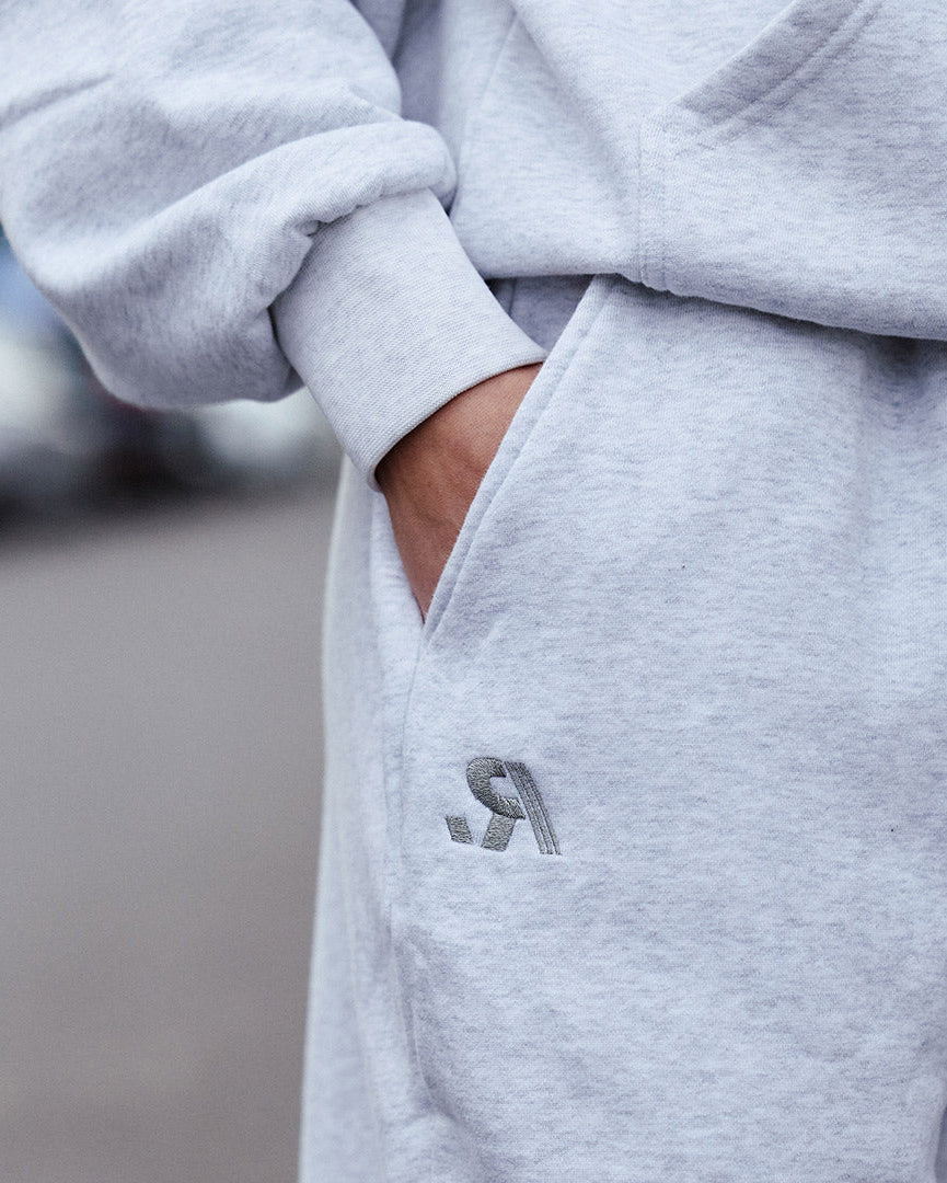 Close-up of a person wearing a light heather gray hoodie and matching sweatpants, with a hand in the pocket and a small dark gray embroidered logo on the thigh.