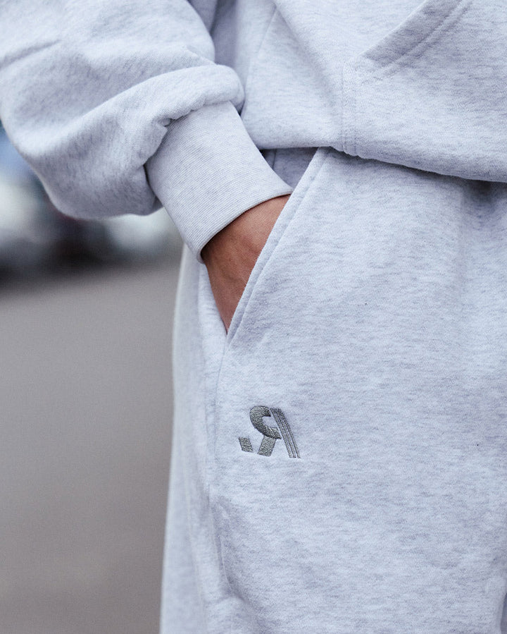 Close-up of a person wearing a light heather gray hoodie and matching sweatpants, with a hand in the pocket and a small dark gray embroidered logo on the thigh.